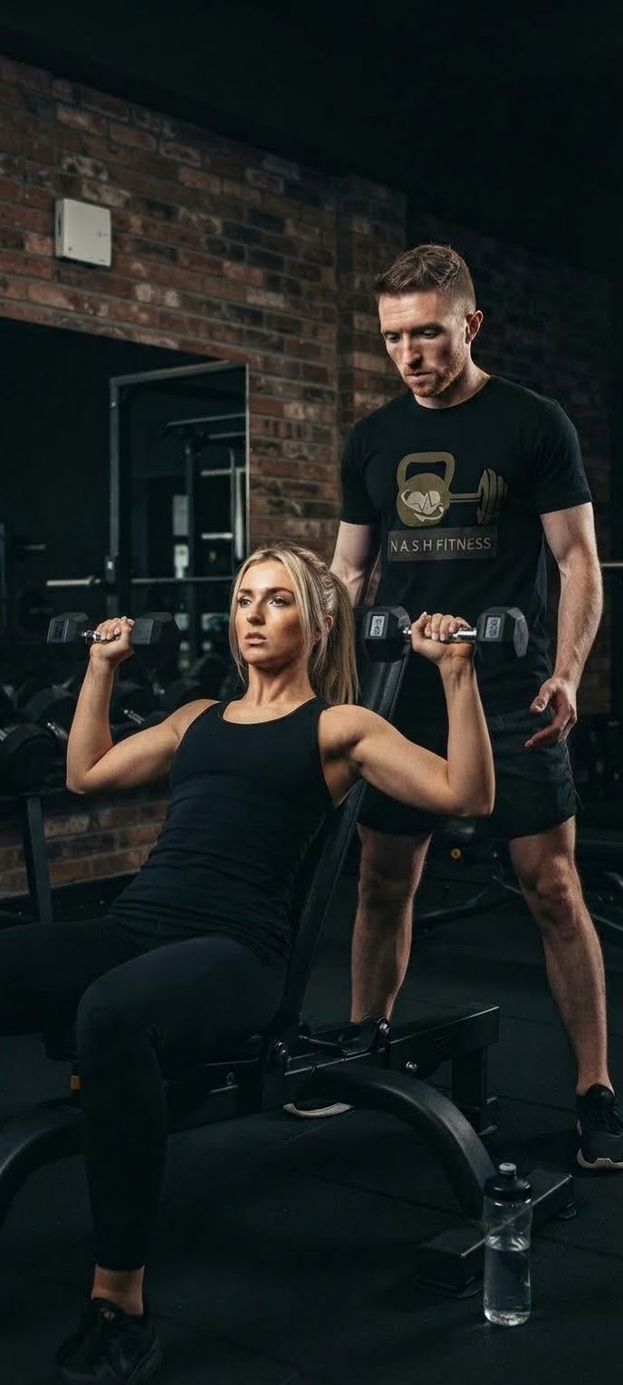 nASH Fitness personal trainer Ashley Crocker spotting a client during an ongoing 1-2-1 dumbbell session, wearing a NASH FITNESS t-shirt
